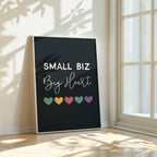 Framed poster with 'Small Biz Big Heart' text and colorful hearts on a black background, placed in a bright room with sunlight casting shadows.