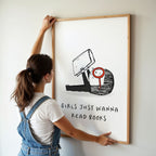 Young woman in coveralls hanging a framed illustration with on a wall. The text in the artwork says 'Girls Just Wanna Read Books.'