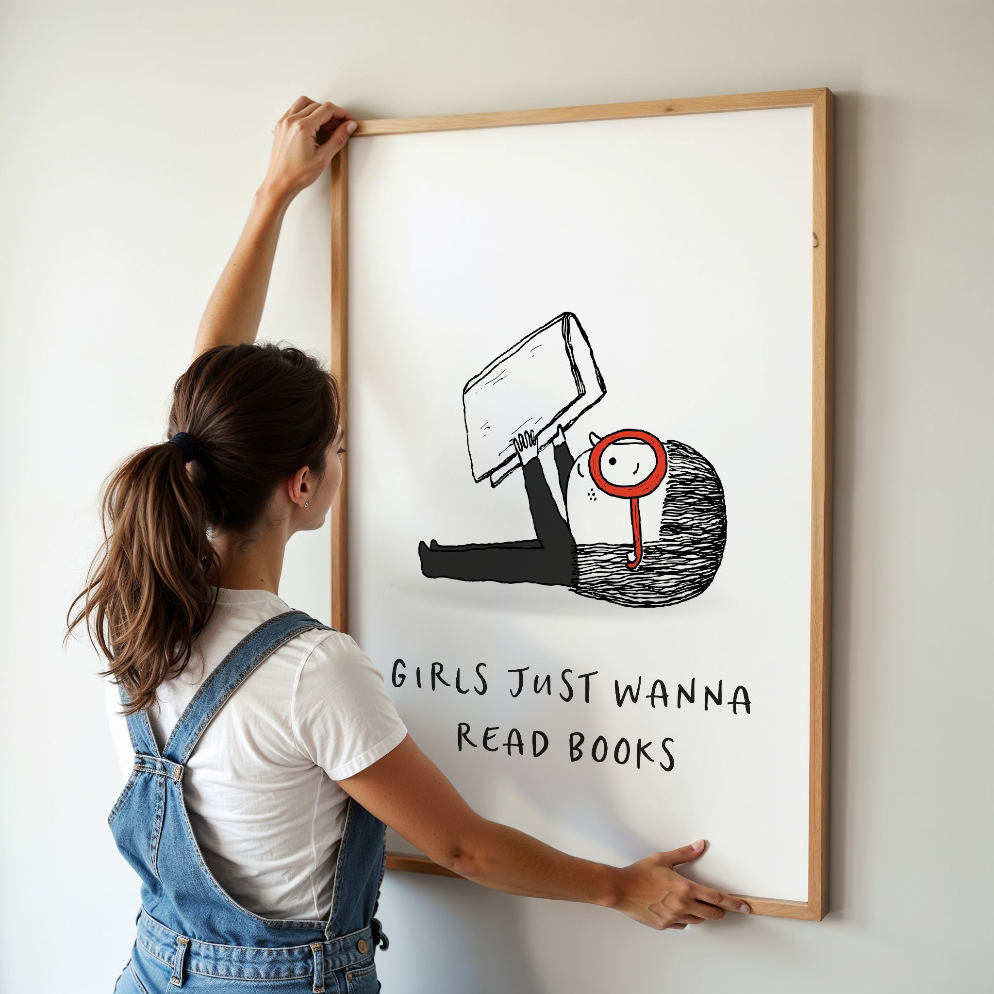 Young woman in coveralls hanging a framed illustration with on a wall. The text in the artwork says 'Girls Just Wanna Read Books.'