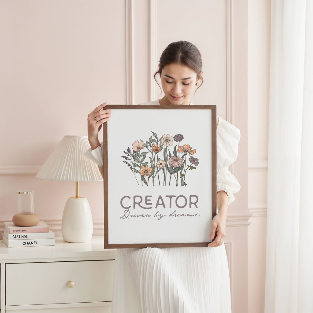 Woman holding a framed artwork with floral design and text in a feminine bedroom with a lamp and books.
