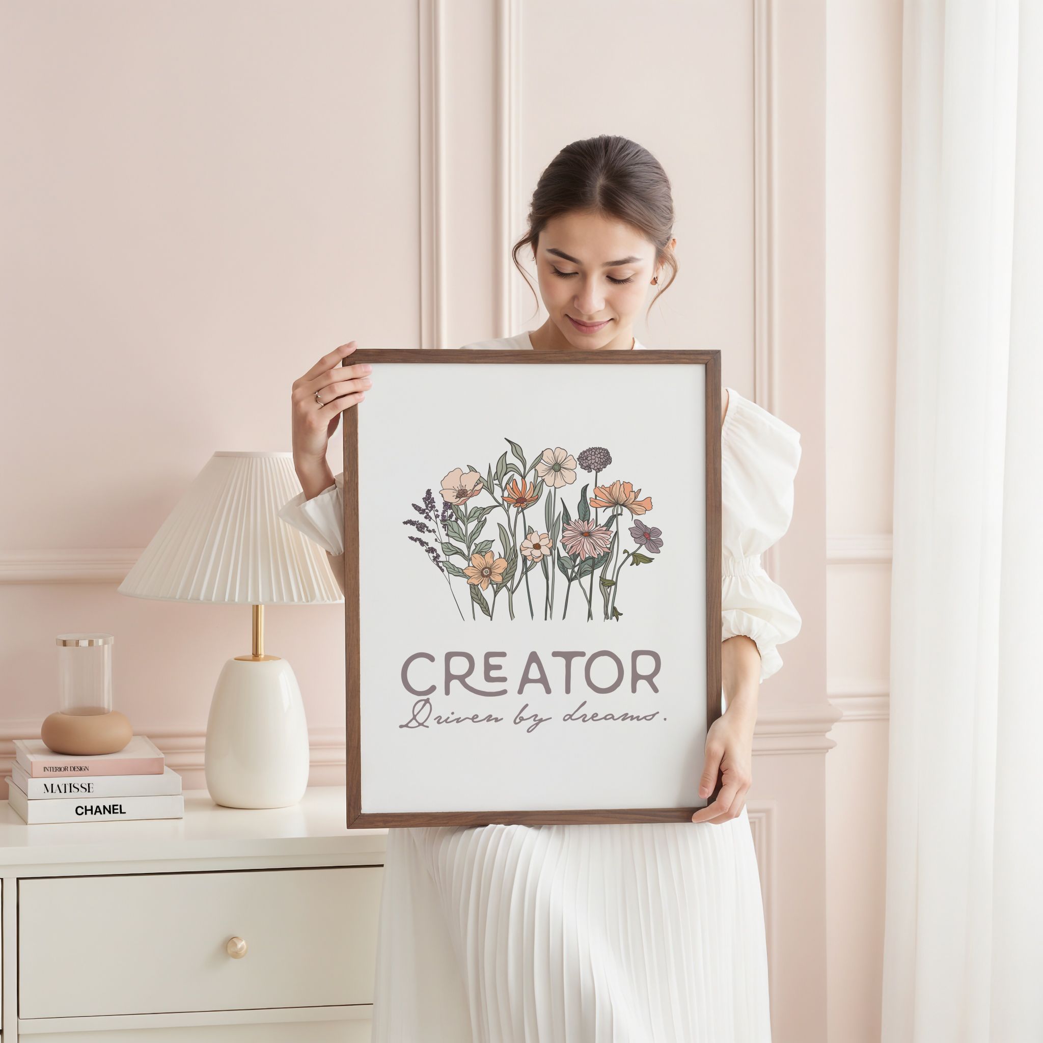 Woman holding a framed artwork with floral design and text in a feminine bedroom with a lamp and books.