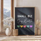 Framed poster with 'Small Biz Big Heart' text and colorful hearts on a wall with a window view of mountains.